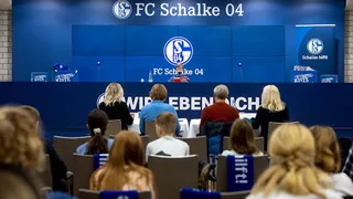 Mehrere Personen, darunter Kinder sitzen auf Stühlen vor dem Pressepult des FC Schalke 04. Vorne an dem Pult sitzt ein Kind mit einem Buch in der Hand.
