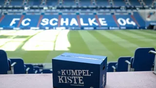 Eine blaue ‚Kumpelkiste‘ mit dem FC-Schalke-04-Logo steht im Stadion vor leeren Rängen auf dem Tisch.