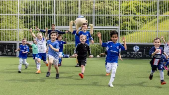 Eine Gruppe lächelnder Kinder in Fußballtrikots rennt freudig mit erhobenen Armen über ein Fußballfeld. Im Hintergrund feuert ein Erwachsener und Erwin das Schalke Maskottchen sie an. Hinter einem Metallzaun ist ein grüner Hügel zu sehen.