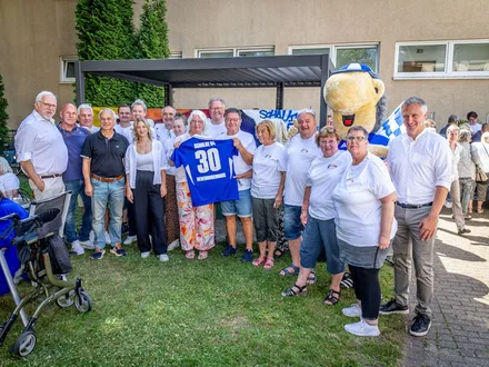 Gruppenfoto bei einer Jubiläumsfeier im Freien. Menschen unterschiedlichen Alters stehen zusammen, in der Mitte wird ein Schalke-Trikot mit der Aufschrift „30 Regenbogenhaus“ hochgehalten. Im Hintergrund ein Maskottchen und weitere Gäste.