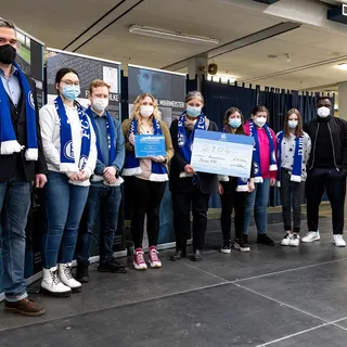 Gruppenfoto in einem Veranstaltungsraum: Eine Gruppe von Schülerinnen, Schülern und Lehrkräften der Gesamtschule Berger Feld präsentiert eine Spende über 2.904 Euro sowie die Ernst Alexander Auszeichnung. Die Personen tragen FC Schalke 04 Schals und Masken. Im Hintergrund sind Infotafeln zur Erinnerungskultur zu sehen.