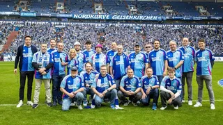 Eine große Gruppe von etwa 25 Personen steht auf dem Rasen der Veltins-Arena in Gelsenkirchen. Alle tragen hellblaue Trikots des FC Schalke 04 mit dem Logo der „Deutschen Krebshilfe“. Die Gruppe steht vor der voll besetzten Nordkurve, deren Fanblock in Blau und Weiß gehalten ist. Die Atmosphäre wirkt energiegeladen und stolz. Ein großes Banner über den Rängen zeigt den Schriftzug „Nordkurve Gelsenkirchen“.