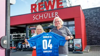 Die Ladenbesitzer halten ein Kumpelpulle-Trikot hoch und lächeln etwas.