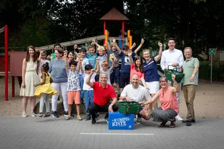Auf einem Spielplatz steht eine große Gruppe aus Kindern und Erwachsenen lachend zusammen. Die Kinder tragen bunte Sommerkleidung und heben begeistert die Arme in die Luft. Im Vordergrund knien mehrere Erwachsene neben einer blauen Kiste mit der Aufschrift „Kumpelkiste“ sowie mehreren grünen Einkaufskörben. Im Hintergrund ist ein Klettergerüst mit Rutsche zu sehen. Die Stimmung ist fröhlich und gemeinschaftlich.