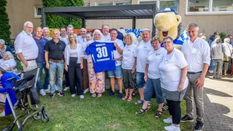 Eine Gruppe von ca. 20 Menschen stehen zusammen. Unter ihnen Schalke Maskottchen Erwin. Sie halten ein Schalke Trikot hoch mit der Nr. 30