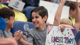 Zwei Jungen in grauen Trikots lächeln und schütteln sich in einem Stadion begeistert die Hände. Einer hält ein buntes Schild mit der Aufschrift „UNSER TEAM“. Im Hintergrund sitzen weitere Kinder mit Schildern.