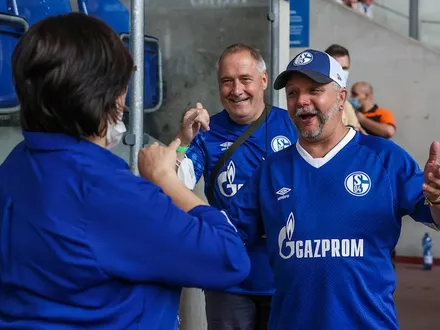 Zwei Männer im Schalke-Trikot strahlen und lächeln eine Dame an und empfangen sie mit offenen Armen.