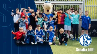 Gruppe von etwa 20 Kindern und Jugendlichen auf einem Fußballplatz, fröhlich posierend mit erhobenen Fäusten, begleitet von Erwin, dem Maskottchen des FC Schalke 04. Viele tragen Fußballtrikots, teils vom FC Schalke 04. Im Hintergrund ein Tor und ein Metallzaun. Links im Bild das großformatige, überlagerte Wort „Schalke hilft!“ mit Vereinslogo.