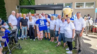Gruppenfoto bei einer Jubiläumsfeier im Freien. Menschen unterschiedlichen Alters stehen zusammen, in der Mitte wird ein Schalke-Trikot mit der Aufschrift „30 Regenbogenhaus“ hochgehalten. Im Hintergrund ein Maskottchen und weitere Gäste.