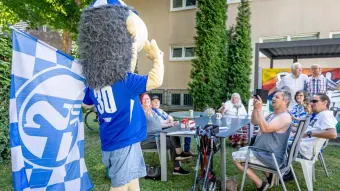 Schalke Maskottchen Erin steht auf einer Wiese vor einem Haus. Auf der Wiese sitzen 4 bis 5 Personen an einem Tisch und schauen fröhlich zu ihm
