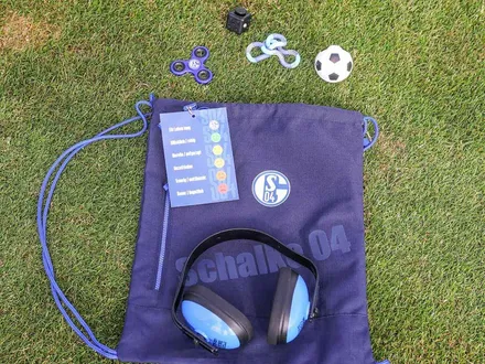 Ein blauer Sportbeutel liegt auf einem Rasen. Zu sehen sind Kopfhörer, ein kleiner Fußball, ein Fidget-Spinner, eine Emotions-Karte, ein kleiner schwarzer Würfel und eine Art Gummidraht.