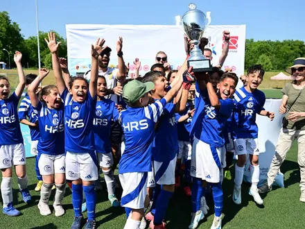 Eine Kinderfußballmannschaft in blauen Trikots des FC Schalke 04 jubelt auf einem Fußballplatz und hält gemeinsam einen großen Pokal in die Höhe. Die Kinder lachen und reißen die Arme in die Luft. Im Hintergrund stehen Erwachsene, die ebenfalls applaudieren und sich mitfreuen. Es ist sonnig und die Stimmung ist ausgelassen und feierlich.