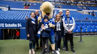 Mehrere Personen, darunter ein Kind, posieren gemeinsam mit dem Schalke-Maskottchen Erwin im Stadion des FC Schalke 04. Alle tragen Schalke-Schals und lächeln in die Kamera. Im Hintergrund sind leere Tribünen und der Spielertunnel mit der Aufschrift „FC Schalke 04“ zu sehen. Die Stimmung ist herzlich und freudig.