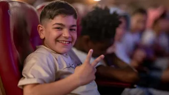 Ein Junge  sitzt in einem Kinosessel und wirkt fröhlich, er zeigt zwei Finger hoch. Im Hintergrund sind weitere Kinder zu erkennen.