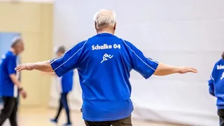 Älterer Mann im Schalke-04-Trikot bei einer Herzsport-Übung in einer Turnhalle, mit ausgestreckten Armen in Balancehaltung.