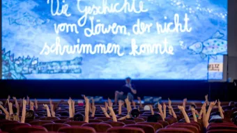 Kinder in einem Kinosaal sitzen auf roten Polstersesseln und strecken begeistert die Hände in die Luft. Auf der großen Leinwand vorne ist in kindlicher Schrift der Titel eines Buches oder Films zu sehen: „Die Geschichte vom Löwen, der nicht schwimmen konnte“. Die Leinwand zeigt außerdem kindlich gezeichnete Fische und ein Krokodil in Blautönen. Eine Person sitzt auf der Bühne vor der Leinwand und spricht vermutlich mit dem Publikum.