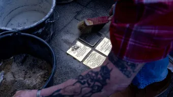 Eine tätowierte Person reinigt mit einer Bürste mehrere Stolpersteine, die in das Pflaster eines Gehwegs eingelassen sind. Neben den Gedenktafeln stehen Eimer mit Sand und Wasser.
