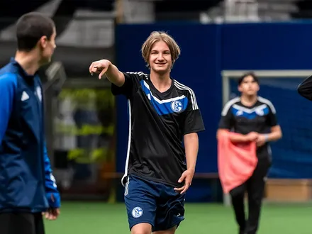 Junge Menschen mit und ohne Behinderung spielen gemeinsam Fußball in einer Indoor-Halle. Ein Spieler im Schalke-Trikot lacht und zeigt auf einen Mitspieler.
