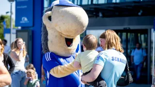 Schalke-Maskottchen Erwin begrüßt lachend ein kleines Kind auf dem Arm einer Frau in Schalke-Trikot beim Familienfest vor der Arena.