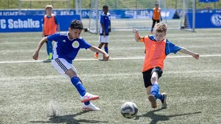 Zwei im blauen Trikot, wovon einer ein orangenes Leibchen trägt laufen auf den Ball zu.