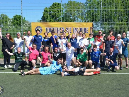 Große Gruppe von Menschen unterschiedlichen Alters und Hintergrunds posiert lachend und jubelnd auf einem Fußballplatz vor einem Banner mit der Aufschrift „Together we inspire“