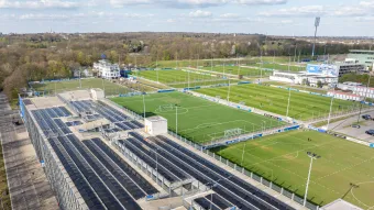 Luftaufnahme des Schalker Vereinsgeländes mit grünen Fußballfeldern, umgeben von Zäunen und Lichtmasten. Angrenzend befindet sich ein Parkhaus mit Solarmodulen auf dem Dach. Im Hintergrund sind Bäume und entfernte Gebäude, wie die Geschäftsstelle und das Rehazentrum zu sehen.