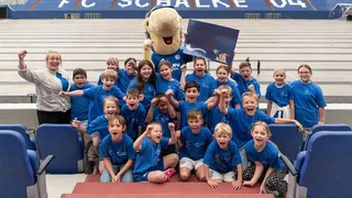 Eine Gruppe von Kindern und ein Erwachsener, alle in blauen Trikots, posieren aufgeregt mit dem Schalke Maskottchen Erwin und einem Schild mit der Aufschrift „Astrid Lindgren Schule“ in einem leeren Stadion, auf dessen Sitzen der FC Schalke 04 zu sehen ist.