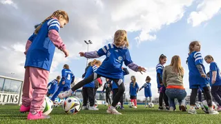 Mehrere junge Mädchen in blauen Trikots mit dem Schalke 04-Logo trainieren auf einem Fußballplatz. Einige kicken Bälle, andere hören einer betreuenden Person zu. Die Szene findet bei Tageslicht unter leicht bewölktem Himmel statt und vermittelt Freude an Bewegung und Gemeinschaft.