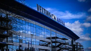 Blick seitlich auf die VELTINS-Arena, der blau-weiße Himmel spiegelt sich in der Glasfront des Stadions