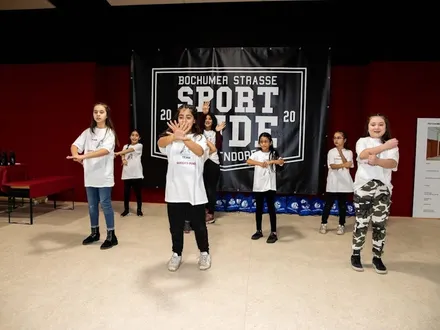 Gruppe von Mädchen in weißen T-Shirts führt gemeinsam eine Tanz- oder Sportchoreografie vor, vor einem Banner mit der Aufschrift ‚Bochumer Straße Sportbude 2020‘.