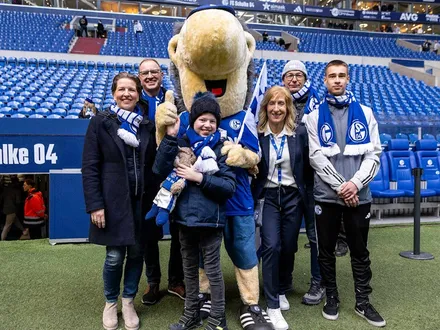 Mehrere Personen, darunter ein Kind, posieren gemeinsam mit dem Schalke-Maskottchen Erwin im Stadion des FC Schalke 04. Alle tragen Schalke-Schals und lächeln in die Kamera. Im Hintergrund sind leere Tribünen und der Spielertunnel mit der Aufschrift „FC Schalke 04“ zu sehen. Die Stimmung ist herzlich und freudig.