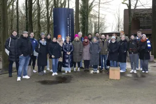 Eine Gruppe von etwa 25 Personen steht eng beisammen vor dem Eingang des Erinnerungszentrums Kamp Westerbork in den Niederlanden. Die Gruppe trägt überwiegend dunkle Winterkleidung mit Mützen und Schals. Im Hintergrund sind kahle Bäume und ein modernes Gebäude zu sehen. In der Mitte des Bildes steht eine hohe, dunkelblaue Stele mit der Aufschrift „Herinneringscentrum Kamp Westerbork“. Die Stimmung ist ruhig und respektvoll.