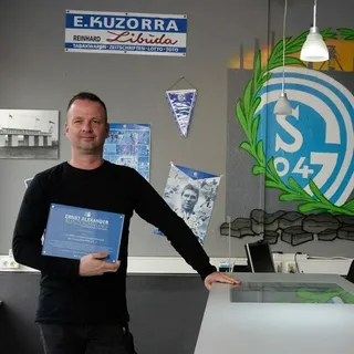 Zwei Männer stehen in einem Raum mit Schalke 04 Fan-Dekoration und halten jeweils eine Tafel mit der Aufschrift "Ernst Alexander Auszeichnung". Im Hintergrund ist ein großes gemaltes Vereinslogo von Schalke 04 sowie historische Fotos und Fanartikel zu sehen.