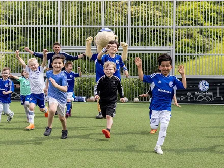 Eine Gruppe lächelnder Kinder in Fußballtrikots rennt freudig mit erhobenen Armen über ein Fußballfeld. Im Hintergrund feuert ein Erwachsener und Erwin das Schalke Maskottchen sie an. Hinter einem Metallzaun ist ein grüner Hügel zu sehen.