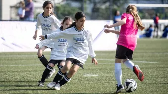 Drei Mädchen im weißen Trikots laufen auf ein Mädchen im pinken Trikot zu, die den Ball am Fuß führt.