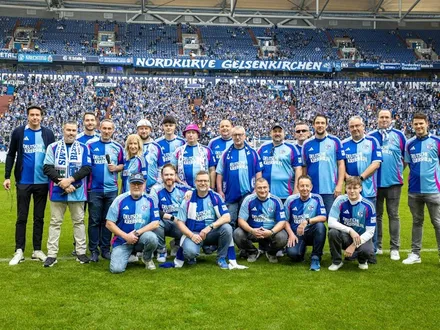 Eine große Gruppe von etwa 25 Personen steht auf dem Rasen der Veltins-Arena in Gelsenkirchen. Alle tragen hellblaue Trikots des FC Schalke 04 mit dem Logo der „Deutschen Krebshilfe“. Die Gruppe steht vor der voll besetzten Nordkurve, deren Fanblock in Blau und Weiß gehalten ist. Die Atmosphäre wirkt energiegeladen und stolz. Ein großes Banner über den Rängen zeigt den Schriftzug „Nordkurve Gelsenkirchen“.