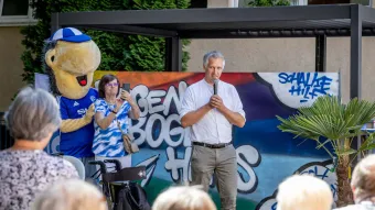 Frank Baumann hält ein Mikro in der Hand und spricht vor einem kleinen Publikum. Im Hintergrund steht das Schalke Maskottchen Erwin.