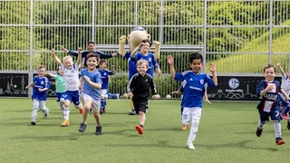 Eine Gruppe lächelnder Kinder in Fußballtrikots rennt freudig mit erhobenen Armen über ein Fußballfeld. Im Hintergrund feuert ein Erwachsener und Erwin das Schalke Maskottchen sie an. Hinter einem Metallzaun ist ein grüner Hügel zu sehen.