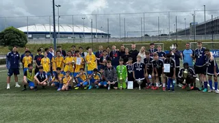 Gruppenfoto von mehreren Jugend-Fußballmannschaften nach einem Turnier auf einem Kunstrasenplatz. Die Spieler tragen Medaillen und präsentieren Pokale und Urkunden. Zwei Mannschaften stehen in der Mitte, eine in gelben Trikots, die andere in dunkelblauen Trikots. Im Hintergrund ist ein großes Stadion mit weißem Dach und ein Parkhaus zu sehen. Der Himmel ist bewölkt. Alle Beteiligten wirken fröhlich und stolz.