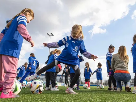 Mehrere junge Mädchen in blauen Trikots mit dem Schalke 04-Logo trainieren auf einem Fußballplatz. Einige kicken Bälle, andere hören einer betreuenden Person zu. Die Szene findet bei Tageslicht unter leicht bewölktem Himmel statt und vermittelt Freude an Bewegung und Gemeinschaft.
