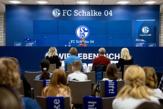 Mehrere Personen, darunter Kinder sitzen auf Stühlen vor dem Pressepult des FC Schalke 04. Vorne an dem Pult sitzt ein Kind mit einem Buch in der Hand.