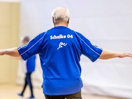 Älterer Mann im Schalke-04-Trikot bei einer Herzsport-Übung in einer Turnhalle, mit ausgestreckten Armen in Balancehaltung.