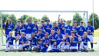 Eine große Gruppe von etwa 25 jubelnden Kindern steht in zwei Reihen vor einem Fußballtor. Alle tragen einheitliche königsblaue Trikots mit dem Logo des FC Schalke 04 und der Aufschrift „HRS“. Die Kinder wirken begeistert, einige reißen die Arme hoch, andere rufen oder lachen. Im Hintergrund ist ein sonniger Tag mit grünem Rasen und Bäumen zu sehen.