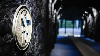 Detailaufnahme des FC Schalke 04-Logos an einer Wand mit dunkler, steinartiger Struktur im Spielertunnel der Veltins-Arena. Im unscharfen Hintergrund ist der Ausgang zum Stadionbereich erkennbar.