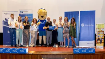 Eine Gruppe von etwa zwölf Personen steht auf einer Bühne und blickt lächelnd in die Kamera, viele zeigen den Daumen nach oben. In der Mitte steht ein Maskottchen im blauen Trikot des FC Schalke 04. Zwei Personen halten eine blaue Kiste mit der Aufschrift „Kumpelkiste“, eine weitere zeigt ein Kinderbuch. Im Hintergrund sind mehrere Roll-Ups mit Logos und Informationen von Unterstützern der Initiative sichtbar, unter anderem von der Stadt Gelsenkirchen, der Kinderbibliothek und dem Arbeitgeberverband Emscher-Lippe. Vor der Bühne stehen weitere „Kumpelkisten“.