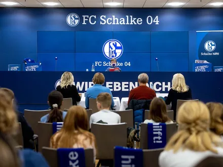 Mehrere Personen, darunter Kinder sitzen auf Stühlen vor dem Pressepult des FC Schalke 04. Vorne an dem Pult sitzt ein Kind mit einem Buch in der Hand.