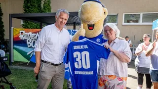 Frank Baumann, Schalke Maskottchen Erwin und eine ältere Dame halten ein Schalke Trikot in die Luft mit der Aufschrift "Schalke 04 30 Regenbogenhaus"