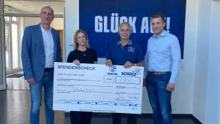 Vier Personen stehen in einem hellen Eingangsbereich vor einer blauen Wand mit dem Schriftzug „Glück Auf“ und halten gemeinsam einen großen Spendenscheck über 8.850 Euro mit dem Empfänger „Schalke hilft!“. Alle lächeln in die Kamera, die Stimmung ist freundlich und professionell.