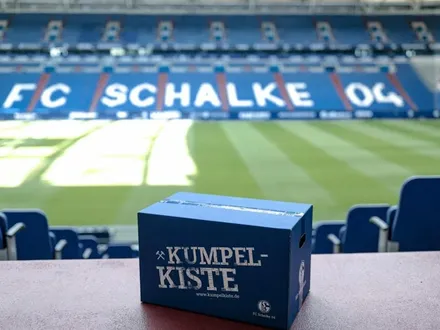 Eine blaue ‚Kumpelkiste‘ mit dem FC-Schalke-04-Logo steht im Stadion vor leeren Rängen auf dem Tisch.