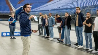 Der Pädagoge erklärt dem Lehrpersonal das Spiel. Sie stehen in der VELTINS-Arena.
